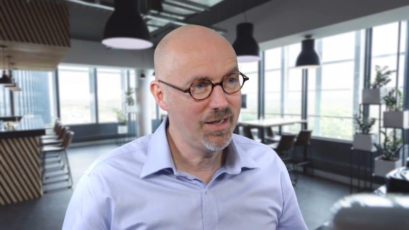 A man with glasses and a light blue shirt looks slightly to the side with a friendly expression. He is indoors, with a modern office background featuring large windows, pendant lights, and chic furnishings.
