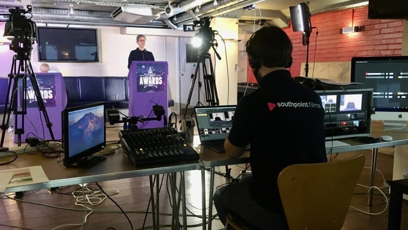 Behind-the-scenes view of a video production setup for the "Staff Awards 2020". In the foreground, a technician with a "southpoint films" shirt operates a video mixer with monitors showing different camera angles. Multiple video cameras are pointed towards a woman at a podium with the awards backdrop. Cables run across the table and floor, emphasizing the complexity of the setup.