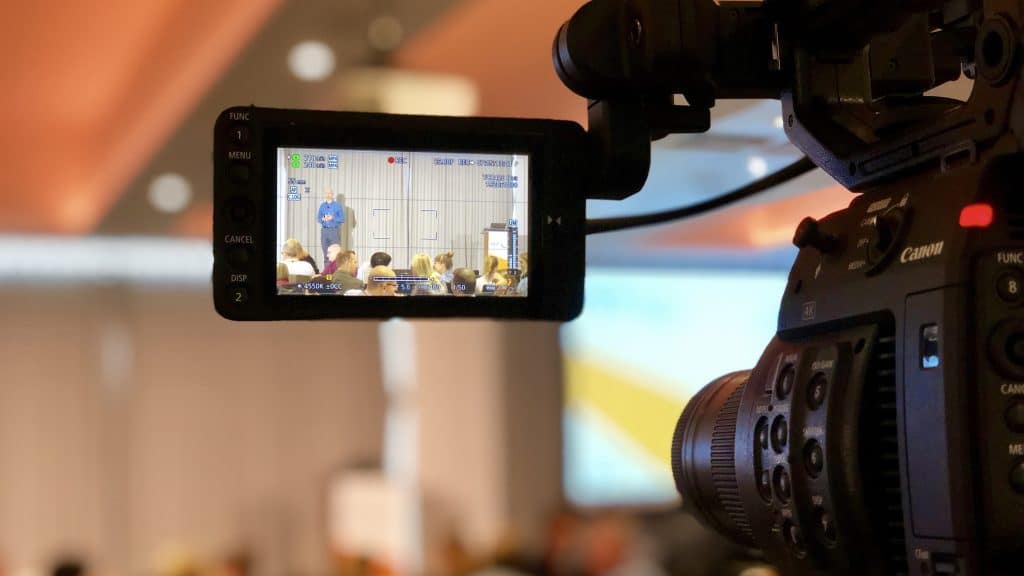 Professional video camera focused on the LCD screen displaying a speaker at a conference. The camera is in sharp focus with details like buttons, brand, and settings visible, while the background, including the audience and the speaker, is out of focus. The perspective gives a behind-the-scenes look at event coverage.
