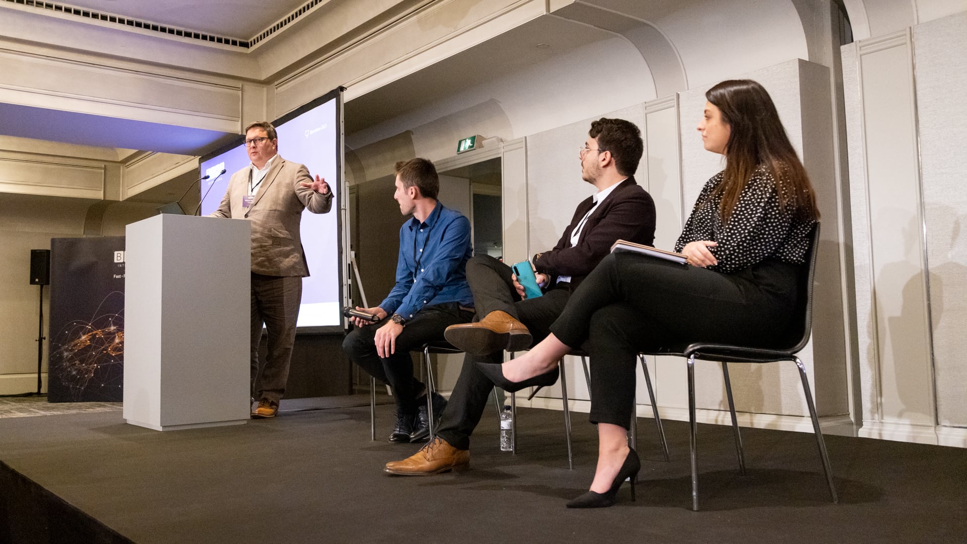 An image from the BOKS Annual Global Conference in Barcelona. A man in a beige suit stands behind a podium, speaking at a conference. Three seated panelists listen attentively; a man in a blue shirt, a man in a dark suit holding a smartphone, and a woman in a black and white polka dot blouse with a notebook. The room has a professional setting with a projection screen and a banner in the background.