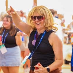 A person with a blonde bob wig and sunglasses is smiling broadly and pointing upwards with their right arm raised, seemingly dancing at an outdoor event. They are wearing a black tank top and lanyards, and other blurred people are in the background.