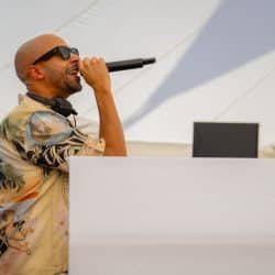 A man with a beard and sunglasses, wearing headphones around his neck, stands behind a white DJ booth, holding a microphone and singing or speaking. He is wearing a patterned short-sleeved shirt with palm trees and cherubs.