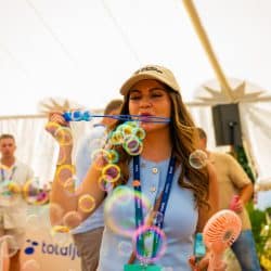 A person with a cap blows bubbles from a blue bubble wand, creating a stream of iridescent bubbles in front of them. They are holding a small pink fan in their other hand. People and a "totaljobs" logo are visible in the background under a white tent.