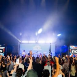 A large crowd of people with their hands raised in the air are at an indoor concert or event, facing a stage lit by blue lights. A band is performing on stage, and two large screens on either side of the stage display the text "noasis". "RECFEST" is visible on a screen above the band.
