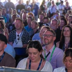 A large crowd of people, many wearing lanyards, are seated and looking forward, seemingly at a stage or speaker. The focus is on the smiling faces of individuals in the foreground, including one person wearing glasses and a lanyard.