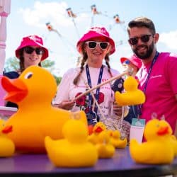 A man with a beard and sunglasses wearing a pink t-shirt smiles at the camera, holding a stick with a small yellow rubber duck attached. Beside him, a woman wearing a pink bucket hat and sunglasses also smiles, holding a similar stick. Numerous yellow rubber ducks are on a purple table in the foreground, and a Ferris wheel is blurred in the background.