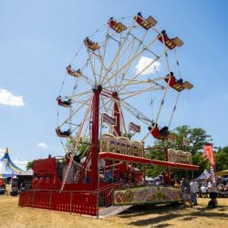 A vintage-style Ferris wheel with red and cream colored spokes and gondolas stands prominently in a sunny outdoor field, surrounded by tents and people at a festival or event.