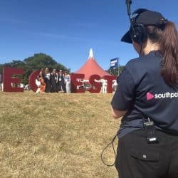 A person wearing a "southpoint films" t-shirt, cap, and headphones stands with their back to the camera, looking towards large red letters spelling "RECFEST" in a grassy field. In the distance, several people are standing, and a red circus-style tent is visible.