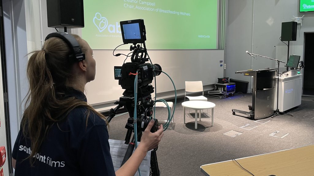 A camera operator, wearing headphones and a "Southpoint Films" shirt, films an empty stage set for a presentation. The stage has a podium with a microphone, a computer, a small white table, and two chairs. A large green presentation slide on the screen reads "ABM - Association of Breastfeeding Mothers" with the presenter's name, Eleanor Campbell, and event hashtag #ABMConf24. The room appears to be prepared for a live or recorded event.