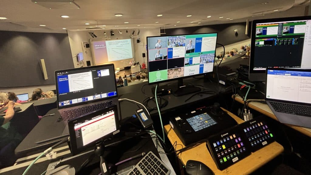 A control booth overlooking a conference or lecture hall. Several screens display video feeds and presentation slides, with control panels and laptops arranged on the desk. The main presentation on the large screen at the front of the room shows a slide titled "What is the opportunity?" with a graph and text. Attendees are seated, and some are using laptops. The setup suggests this is a live event being managed or streamed.