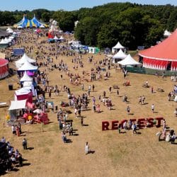 An aerial view of a large outdoor festival or event, with numerous colorful tents, including two prominent red and white circus-style tents. Many people are scattered across a large grassy field, and a large "RECFEST" sign is on the ground. Trees line the background.