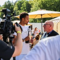 A man with curly hair, wearing a white t-shirt and holding a microphone with a "RECFEST" logo, is interviewing another man wearing glasses and a dark t-shirt. A video camera and a cameraman are partially visible in the left foreground, and a yellow umbrella and other people are in the sunny background of an outdoor event.