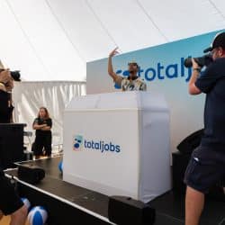 Two photographers, one on the left in black and one on the right in a "southpoint films" shirt, are actively shooting a DJ on a stage. The DJ, wearing sunglasses and a patterned shirt, stands behind a white "totaljobs" branded booth, raising one arm. A large "totaljobs" banner is in the background.
