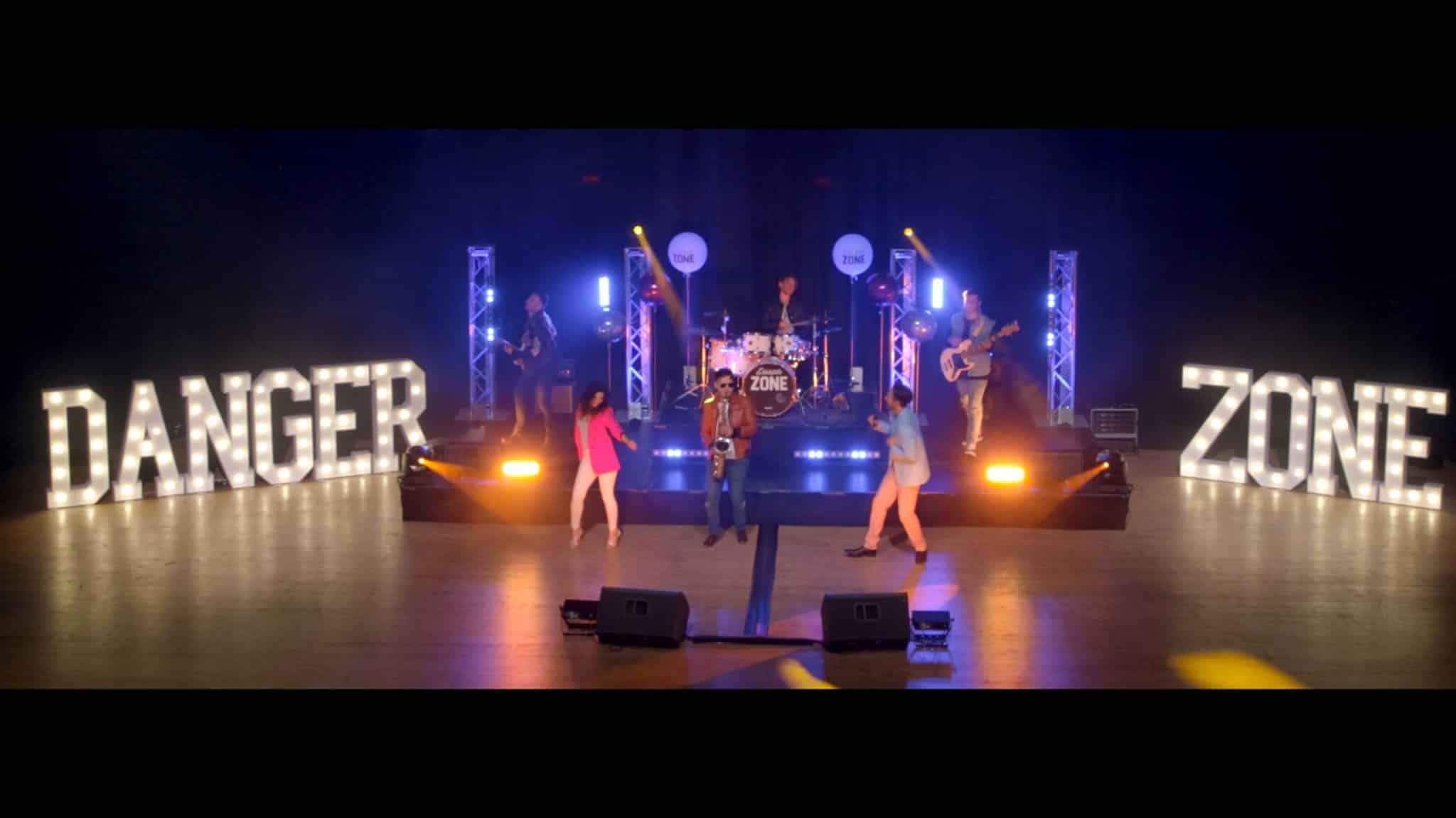 A live music performance on a brightly lit stage featuring a band. Large illuminated letters spelling "DANGER" and "ZONE" flank the stage. The band includes a drummer, guitarist, and bassist, and saxophonist. Two singers in colourful outfits perform in front of the band. The stage is lit with vibrant blue, purple, and yellow lighting, adding to the energetic atmosphere.