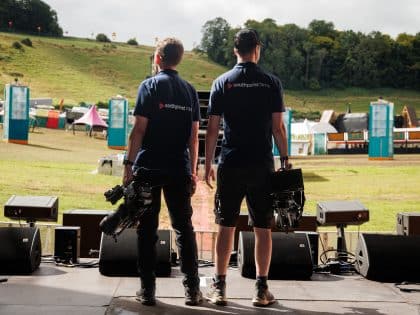 Two videographers, seen from behind, stand on an outdoor stage, each holding professional camera equipment. They are wearing matching navy blue shirts with "Southpoint Films" printed on the back. In the background is a grassy hillside with colorful tents, festival booths, and speaker towers, suggesting they are at a festival or event. The lighting and shadows indicate late afternoon, and the scene captures a calm, pre-event atmosphere.