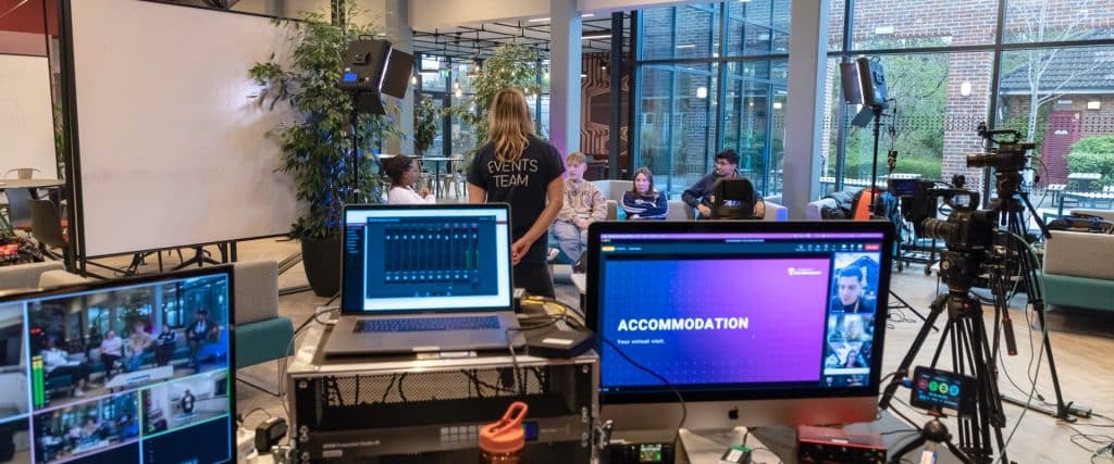 A video production studio setup with cameras, lights and video streaming equipment on show. The venue is a university common room and there are a panel of students on sofas. The cameras are pointed at them. They are hosting a virtual event about university accommodation, as indicated by the content on the computer screens.