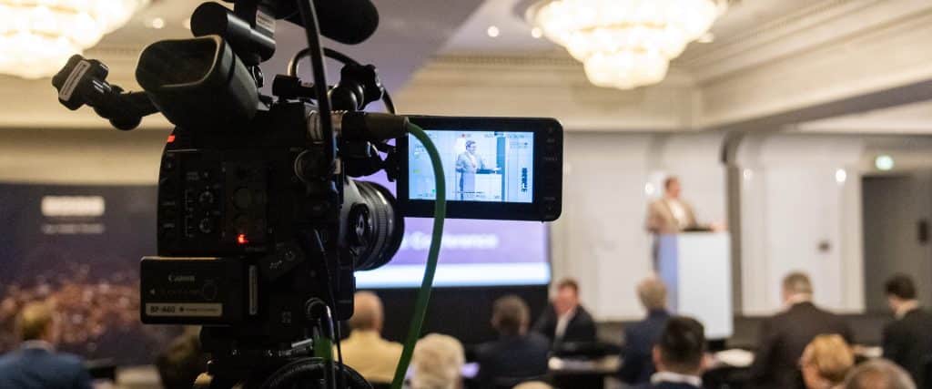 A man stands at a lectern in a conference centre. The crowd can be seen in front of him. In the foreground, a professional video camera is seen recording him.