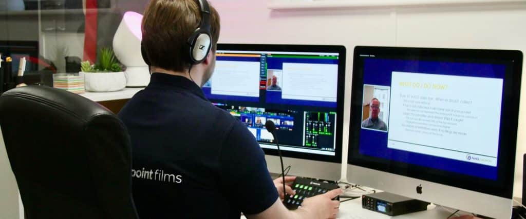 A man wearing a polo shirt that reads "Southpoint Films" on the back is sat a desk. There are two large computer monitors in front of him and he is holding a control panel with lots of buttons. On the left-hand screen is video streaming software vMix. On the right-hand screen is a large blue screen with a "picture in picture" of the presenter and their slides. The presenter is using a webcam.