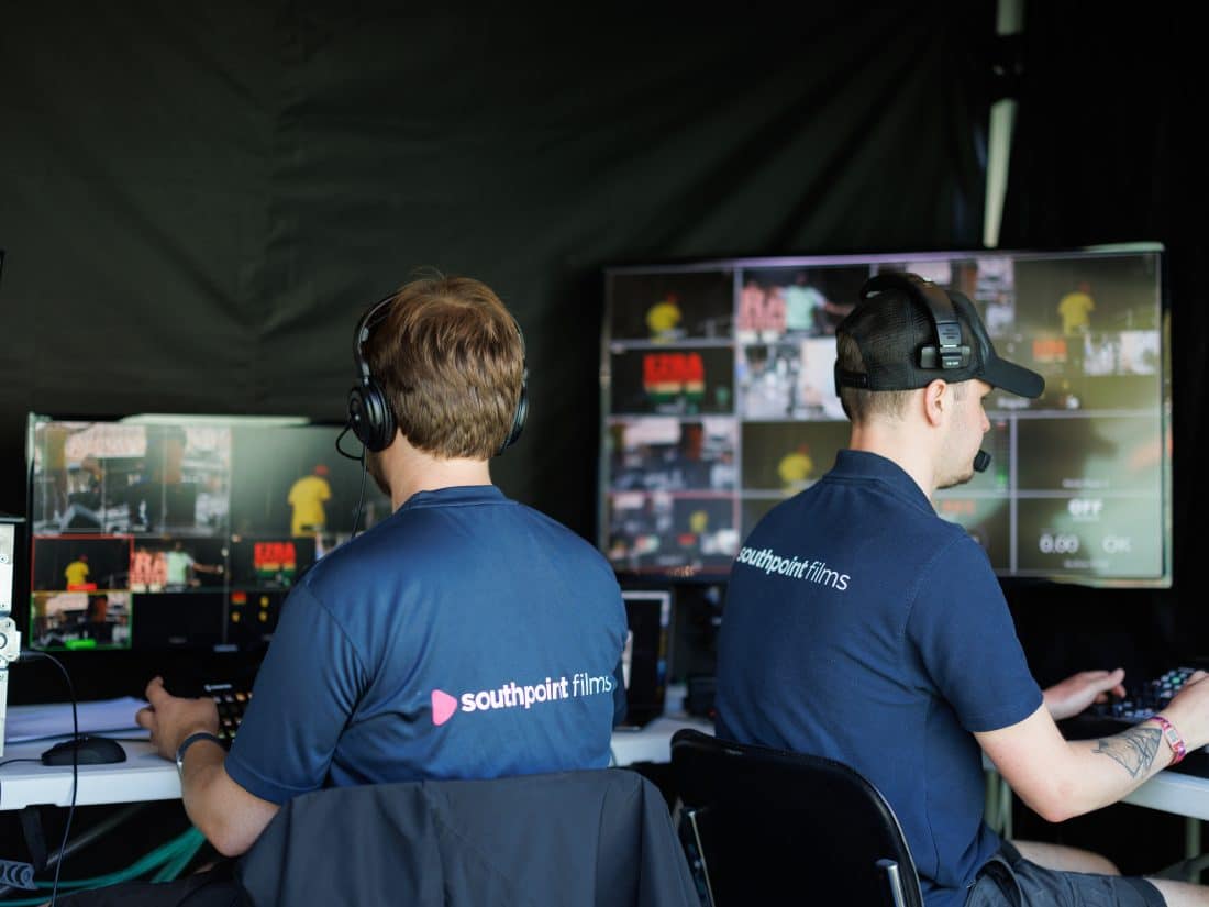 Two crew members wearing headsets and navy blue "Southpoint Films" shirts sit at a table in front of multiple monitors displaying live video feeds. One person operates a switcher while the other monitors the screens. The workspace includes technical equipment, cables, and a dark backdrop.