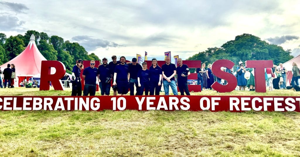 A group of eight people stand in front of large red letters spelling "REC FEST" with a banner below reading "CELEBRATING 10 YEARS OF RECFEST." The scene is outdoors with a grassy field, a red tent, and trees in the background. Several people are walking and mingling in the area, indicating a festival or event atmosphere