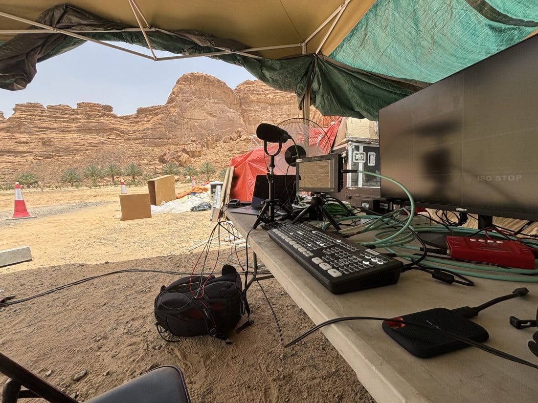 A mobile broadcasting setup is seen under a canopy in the Saudi Arabian desert. The equipment includes a laptop, microphone, large monitor displaying a program labeled "Rami Kadi," video switcher, and various cables. A rocky landscape with cliffs, palm trees, and red traffic cones is visible in the background. A fan, bags, and orange equipment are scattered nearby, indicating a temporary outdoor setup.