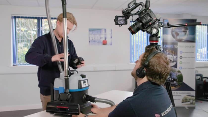 A video production setup in an office environment. A person in a navy blue sweater works on a cleaning machine, while another person wearing headphones operates a professional video camera on a tripod. The scene is brightly lit with windows in the background and a banner reading "Dare to Innovate" visible on the right. The cleaning machine is branded with "ICE".
