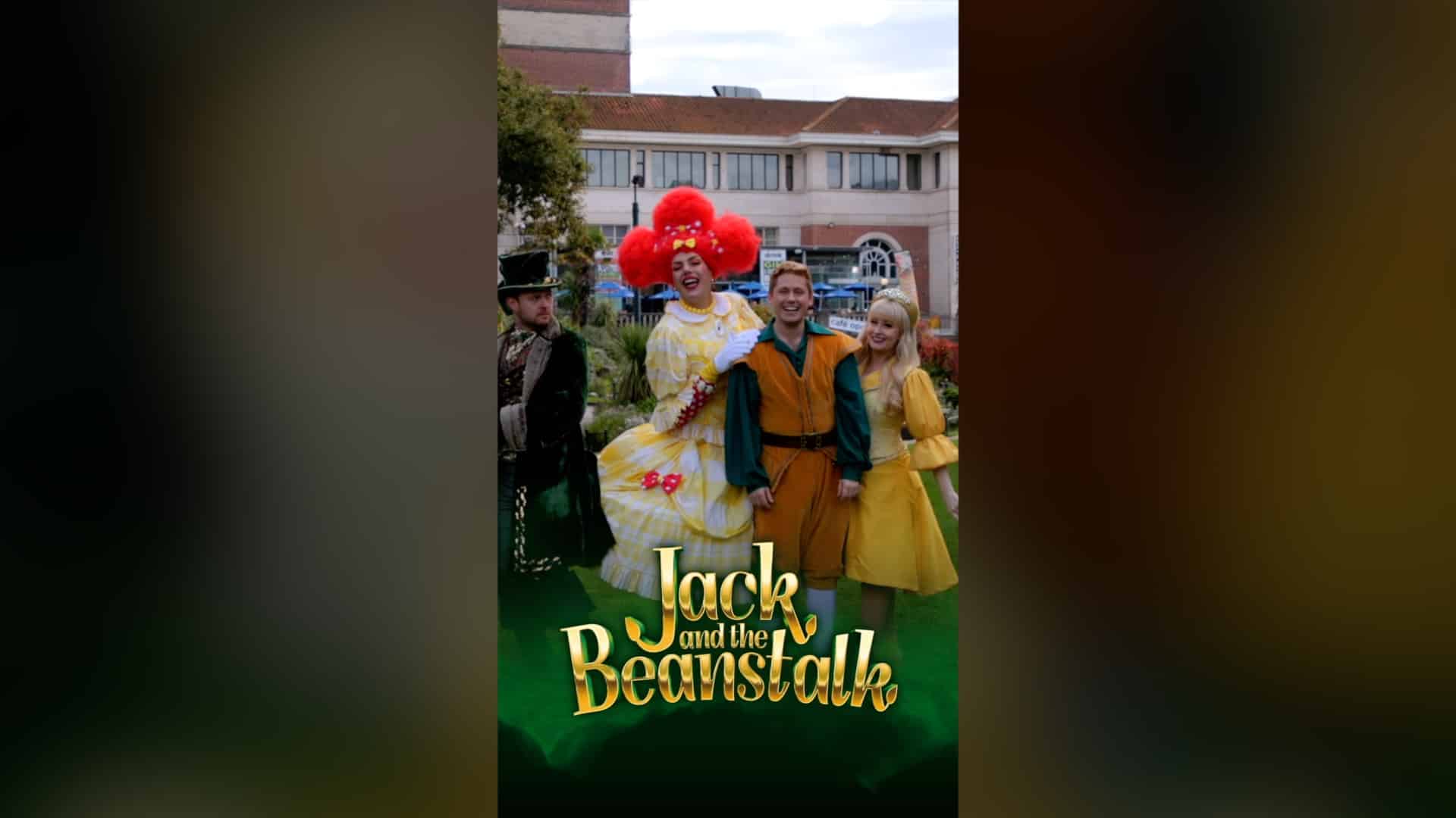 Four actors in colorful costumes pose outdoors for a promotional image of Jack and the Beanstalk. The central figure, dressed in a bright yellow dress with a large red wig, appears to be a pantomime dame, laughing while holding the arm of a man dressed as Jack in an orange and green outfit. To Jack’s right, a woman in a golden princess-style dress smiles, while on the left, a man in a dark green and black costume with a top hat looks on suspiciously. The title Jack and the Beanstalk is displayed in bold golden letters at the bottom of the image. The background features a public square with a red-brick building.
