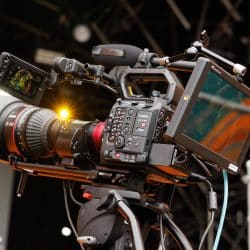 A professional Canon EOS C400 video camera is mounted on a tripod, equipped with a large zoom lens, an external monitor, and various attachments. The camera is set up on a stage with lighting and rigging in the background, capturing a live event. A bright lens flare reflects off the camera, adding a dramatic effect.
