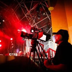 A videographer operates a professional camera on a tripod, capturing a live performance bathed in intense red stage lighting. The operator wears a headset and is positioned near the side of the stage, surrounded by metal rigging, lights, and equipment. The dramatic lighting and shadows highlight the energy of the event.