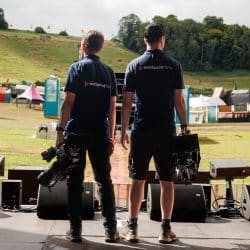 Two videographers stand on a stage, facing an outdoor festival setup with tents, stalls, and a grassy hillside in the background. They wear matching navy-blue shirts with "southpoint films" printed on the back and hold professional video cameras. Large speakers line the stage, and the scene is bathed in natural sunlight.