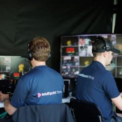 Two video production crew members from "Southpoint Films" sit at a control desk inside a tent, monitoring multiple screens displaying a live event. They wear matching navy-blue shirts with the company logo and headsets for communication. One operates a video switcher while the other observes the footage. The workspace is equipped with monitors, cables, and professional broadcasting equipment.