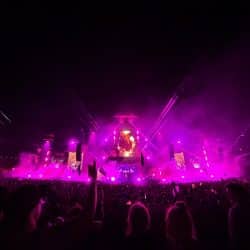 A massive crowd enjoys a nighttime music festival, illuminated by vibrant pink and purple stage lighting. Beams of light and smoke effects create a dramatic atmosphere as a large screen displays visuals of the performer. Festivalgoers raise their hands and wave flags, adding to the energetic scene.