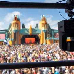 A mounted remote-controlled camera overlooks a large festival crowd in front of a colorful, ornate stage with tall towers and an arched centerpiece. The bright daytime sky contrasts with the vibrant architecture, while festivalgoers wave flags and enjoy the event. The camera setup suggests professional video production capturing the scene.