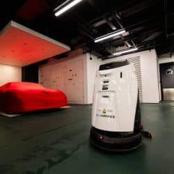 A white autonomous floor-cleaning robot in a modern underground garage, with a red car fully covered by a fitted cover featuring a black Ferrari logo. The space is lit by overhead industrial lights and features dark flooring, white walls, and a metallic double door in the background.