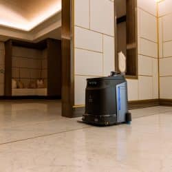 A black autonomous cleaning robot moving across a polished marble floor in an upscale interior with modern, gold-accented wall panels. The space includes a recessed seating area with beige cushions, soft ambient lighting, and a clean, minimalistic design.