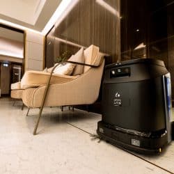 A sleek autonomous cleaning robot on a polished marble floor in a luxurious indoor setting. The space features modern beige seating with metal frames, dark wood paneling, and warm ambient lighting. In the background, decorative plants and glass doors add to the upscale interior design.