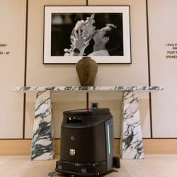 A compact autonomous cleaning robot stationed on a wooden floor beneath a marble console table with a ceramic vase. Above the table hangs a black-and-white framed photo of a tennis player kissing a trophy. The scene is set in a sleek, modern interior with neutral tones and minimalist decor.
