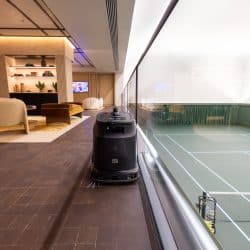 An autonomous floor cleaning robot positioned in a modern indoor lounge area with a view into a glass-enclosed tennis court. The lounge features stylish furniture, warm lighting, and a wall-mounted TV, while the tennis court is clean and well-lit with crisp white lines on the green surface.