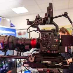 A professional Canon EOS C400 cinema camera mounted on a tripod rig with a large Canon 17-120mm lens attached. The camera is equipped with various controls and buttons on its side panel and a top handle with an attached monitor. It is positioned indoors, likely at a tech or media event, with people and display monitors visible in the blurred background.