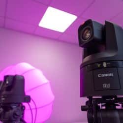 A close-up of a Canon 4K PTZ (pan-tilt-zoom) camera mounted on a tripod in a studio setting with soft pink lighting. Another similar camera is visible in the background next to a large glowing pink light diffuser. The ceiling and walls of the studio are also bathed in pink light, creating a vibrant and modern atmosphere.