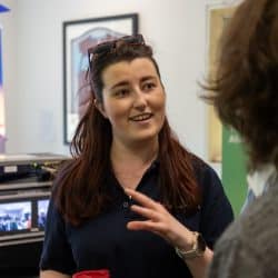 A woman with long hair, wearing a navy polo shirt and sunglasses on her head, is engaged in conversation at an event. She is smiling and gesturing with her hand while holding a red cup. In the background, there is technical equipment with monitor screens, a banner and a framed picture on the wall. Another person is partially visible in the foreground, facing her.