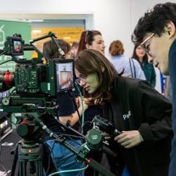 Two individuals closely examine a professional Canon EOS C400 cinema camera setup on a tripod, with a large Canon 17–120mm lens and an external monitor showing a live feed. One person, wearing a jacket with 'Pav' on it, is looking into the camera's monitor while holding the tripod's handle. The other leans in from the right side. In the background, more people are gathered, and a green banner with a white media icon and text is visible, indicating a media or broadcast-related event.