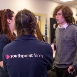 A group of people engaged in conversation at an indoor event. In the foreground, a person wearing a navy blue 'southpoint films' polo shirt is seen from the back, talking to a man in a grey sweater with long, curly hair. Another woman with long hair, also in a 'southpoint films' shirt, is partially visible beside them. The background includes more attendees and a table with promotional materials, suggesting a networking or industry event
