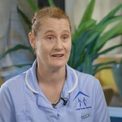 A woman in a light blue care worker uniform sits indoors, speaking during what appears to be an interview. Her uniform displays the logo and name 'Lodge Group Care UK.' She has light hair pulled back, and is wearing a small microphone. The background features plants, soft furnishings, and natural light.