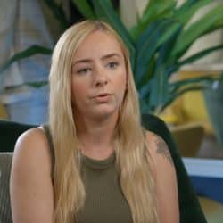 A woman with long blonde hair sits indoors, speaking during an interview. She is wearing a sleeveless green top and has a visible tattoo on her upper arm. A microphone is clipped to her clothing. The background includes a green plant, cushioned seating, and soft natural lighting.