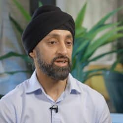 A man wearing a black turban and a light blue shirt speaks during an interview. He has a beard and is wearing a clip-on microphone. The background includes indoor plants, curtains, and cushioned furniture, creating a warm and inviting setting.