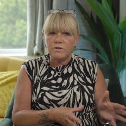 A woman with blonde hair and bangs sits indoors during an interview, wearing a sleeveless black and white patterned top. She has glasses resting on her head and is speaking with her hands slightly raised. A tattoo is visible on her forearm. The background includes a yellow armchair, a green plant, and a window letting in natural light.