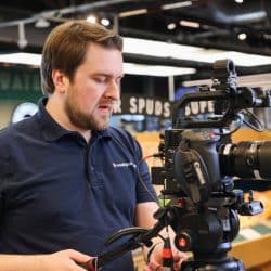 Man in a navy blue “Southpoint Films” shirt operating a Canon camera inside a grocery store. He appears focused on adjusting the camera, with shelves of produce in the background.