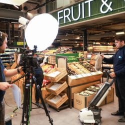 Film crew shooting a video inside a grocery store’s produce section. A camera operator films a man using a floor cleaning machine while another crew member holds a boom mic. Bright studio lighting illuminates the set.
