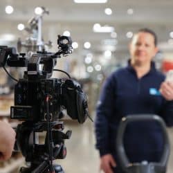 Close-up of a professional Canon video camera on a tripod, filming a woman in the background who is holding a phone and operating a floor cleaning machine in a store.